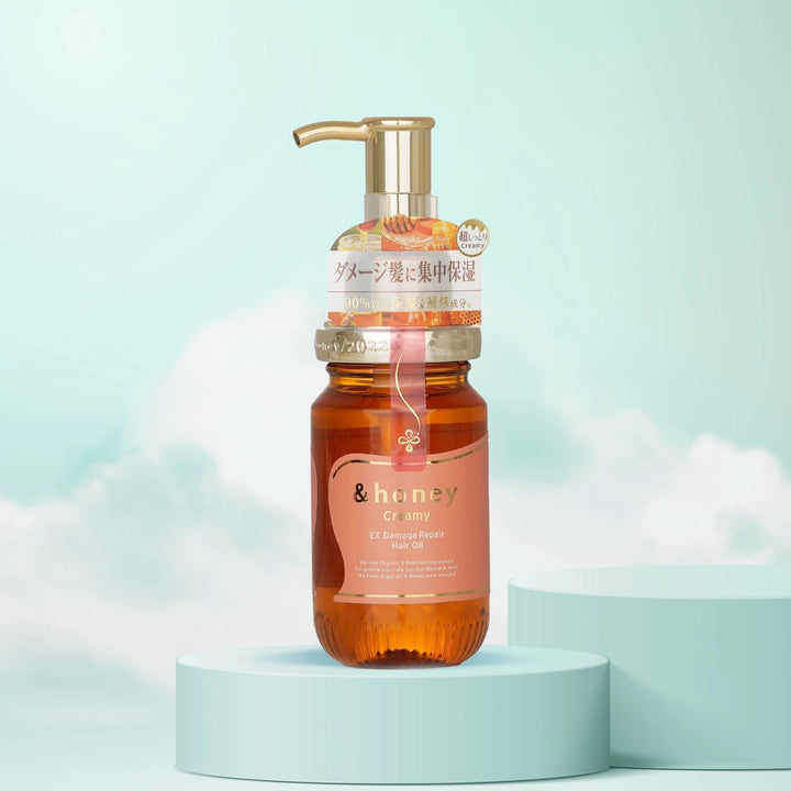 A bottle of &Honey Creamy EX Damage Repair Hair Oil with a pump dispenser, placed on a cylindrical pedestal against a sky blue background with clouds.