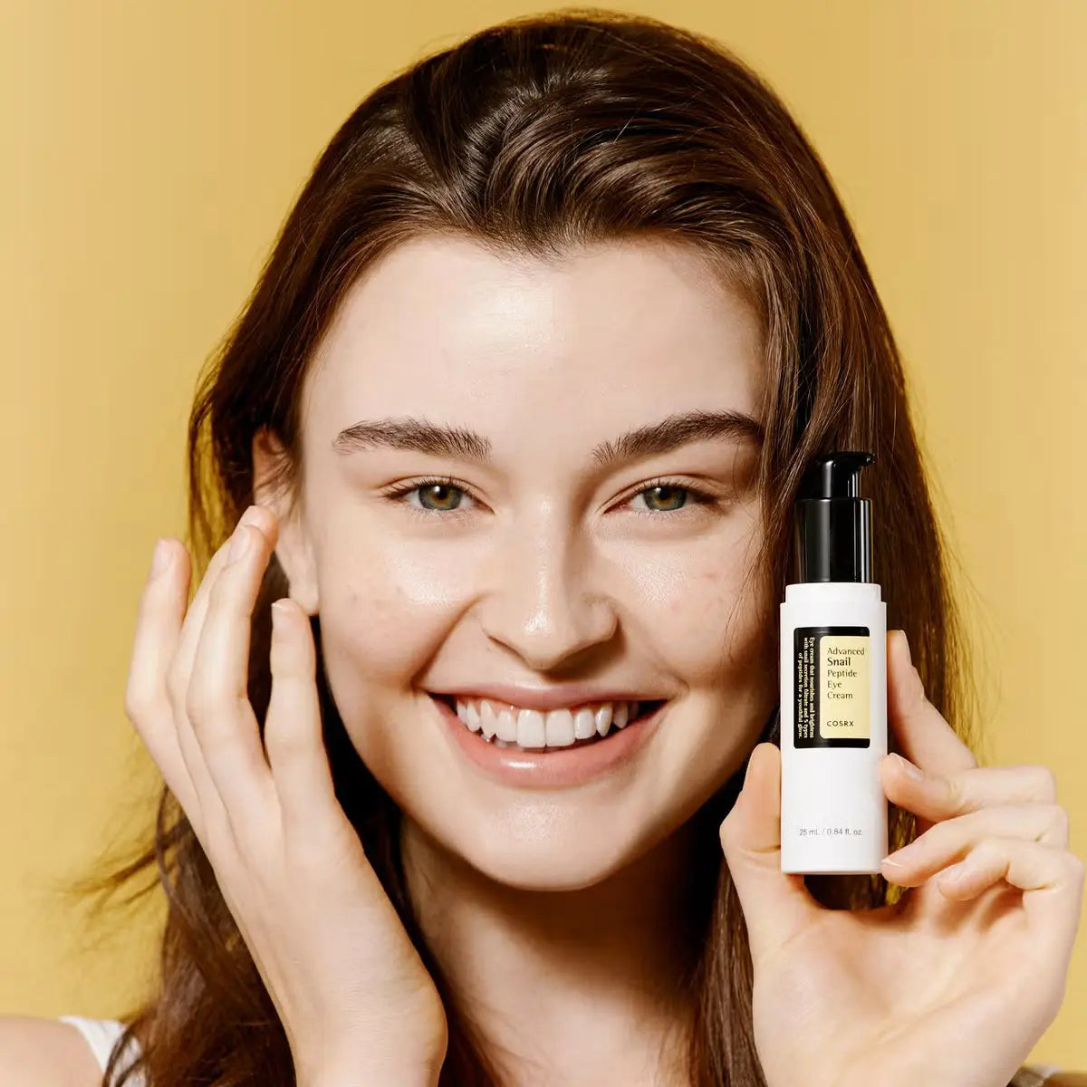 Woman holding a Cosrx Advanced Snail Peptide Eye Cream bottle against a yellow background