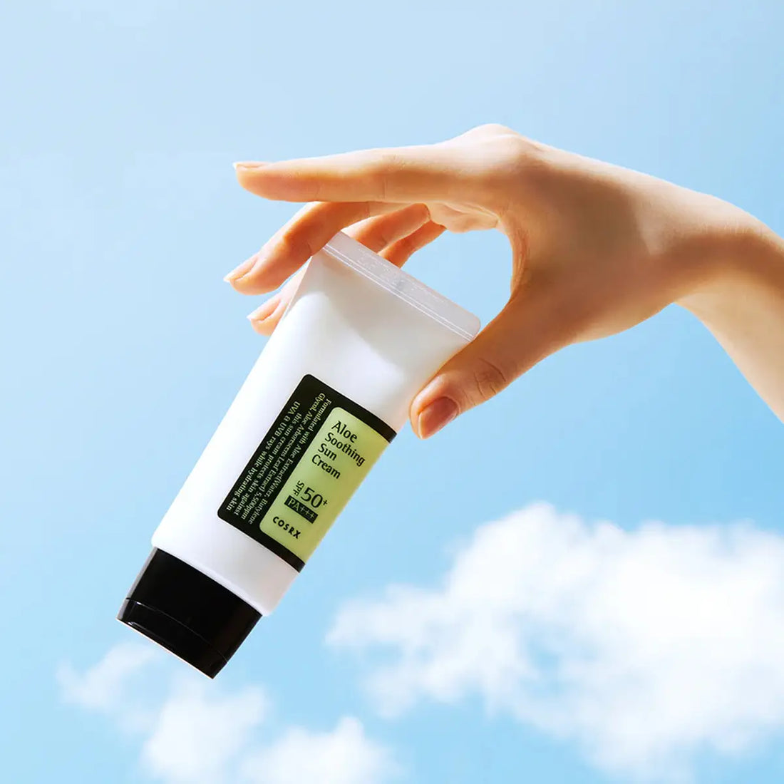 Hand holding a Cosrx Aloe Soothing Sun Cream with a blue sky background