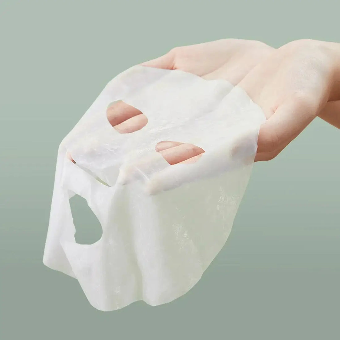 Hand holding a white cube-shaped mask with cutouts on a light green background