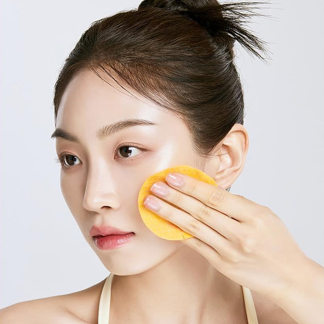 Woman applying a yellow facial product with a sponge on a plain background
