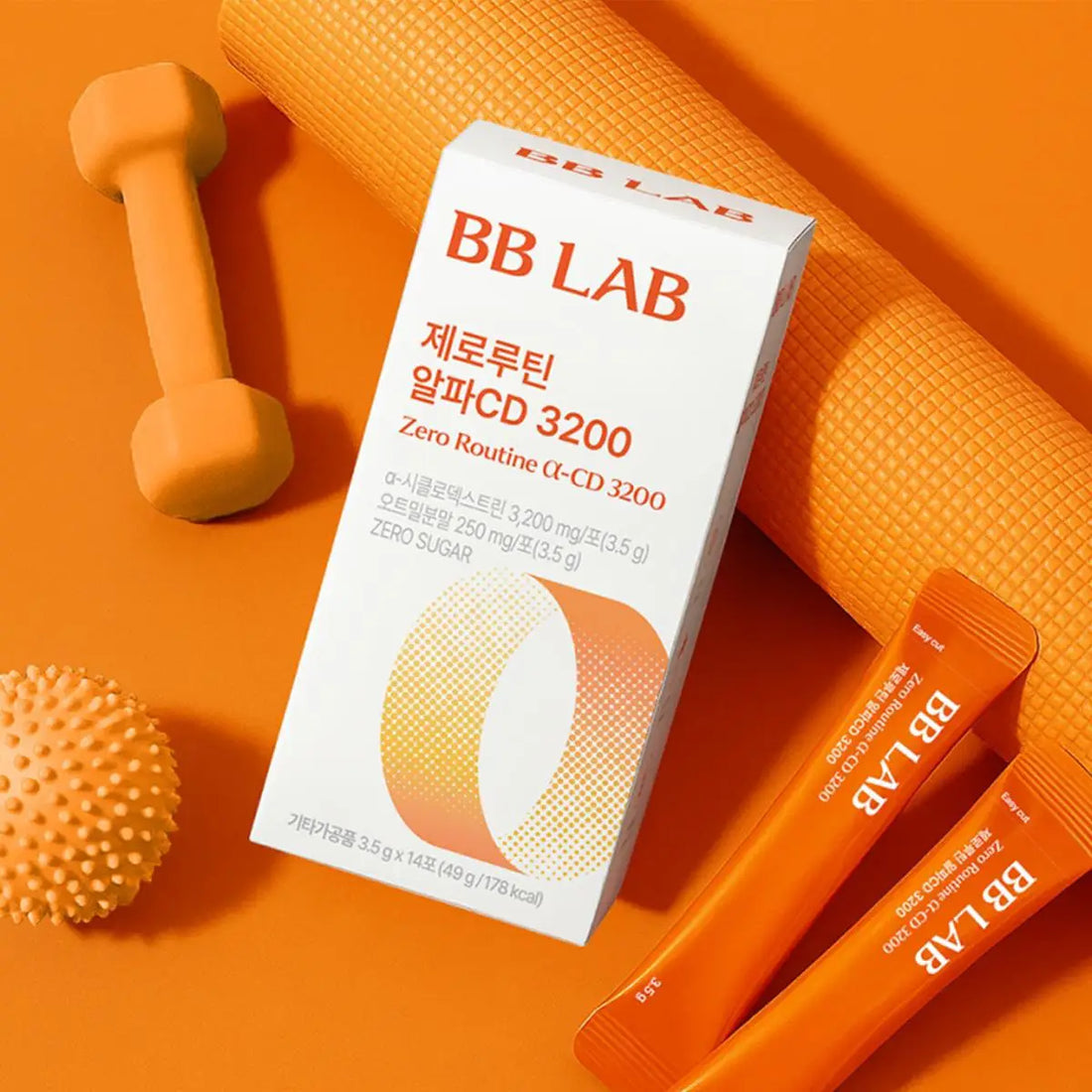 BB LAB Zero Routine (α-CD) 3200 supplement packaging, including a white box with orange text and two orange single-use sachets, styled on an orange background with a dumbbell, textured ball, and yoga mat.