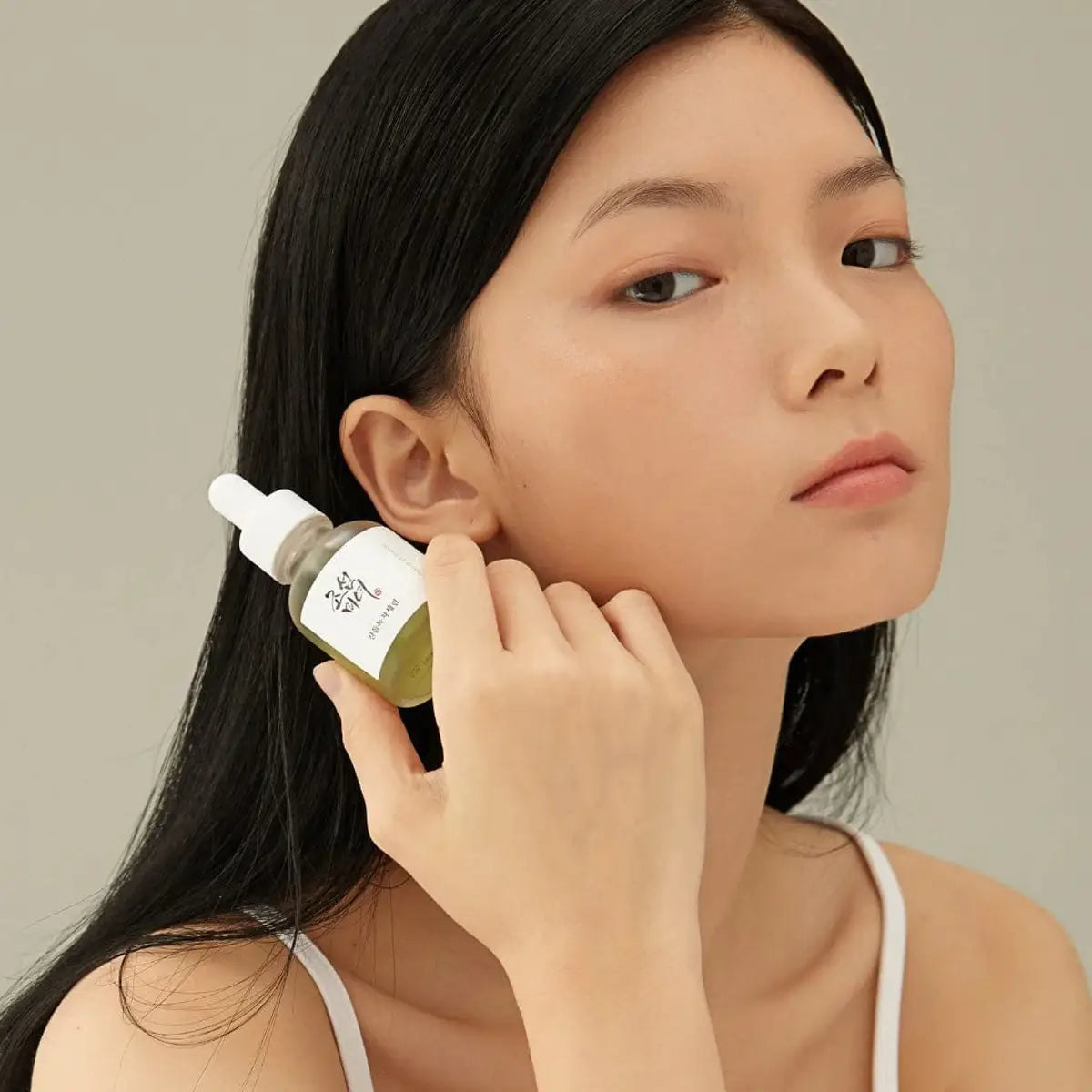 Woman holding a dropper bottle near her ear against a neutral background