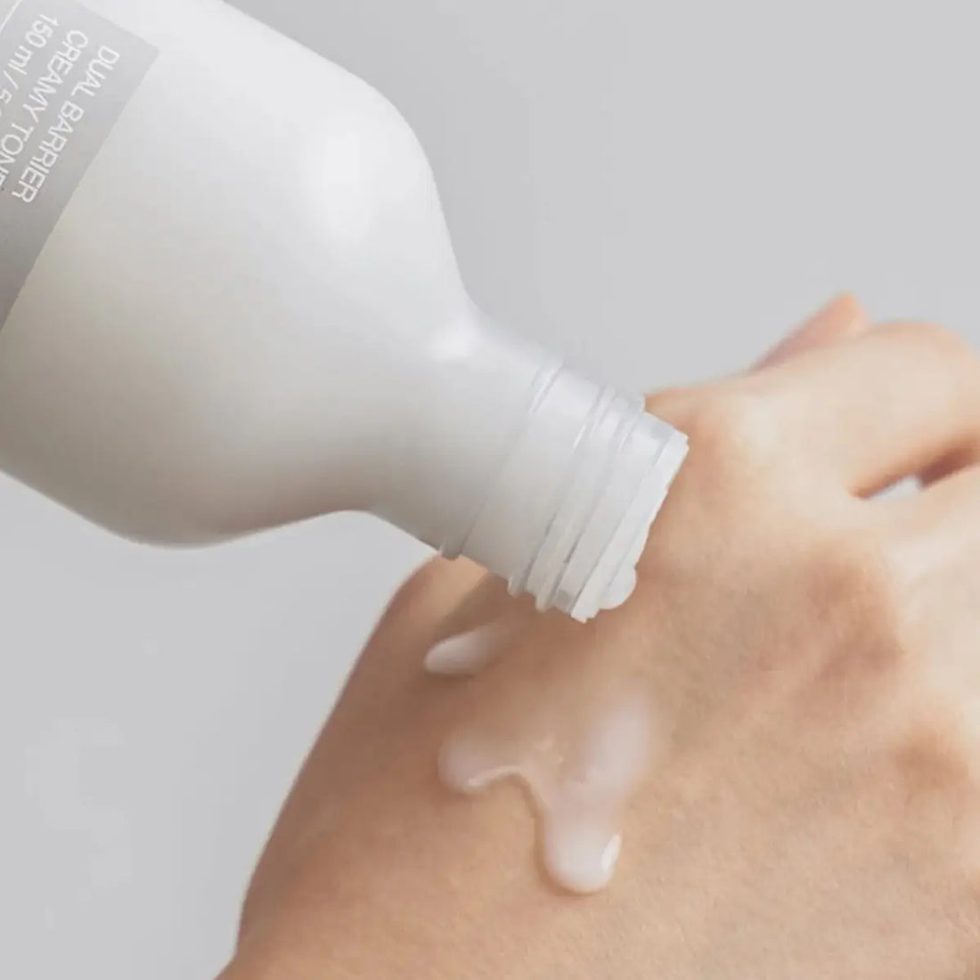 Hand applying a white cream from a bottle onto skin against a light gray background