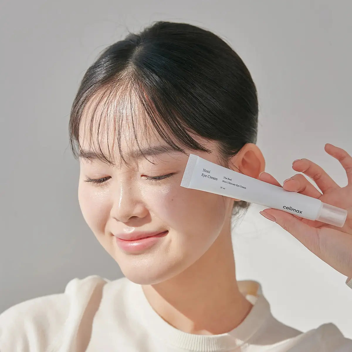Woman holding a tube of Cellmax eye cream against her face on a plain background