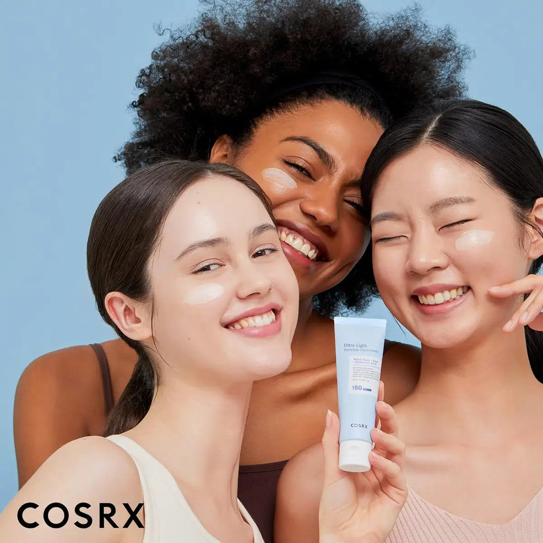 Three women smiling and holding a COSRX skincare product against a blue background