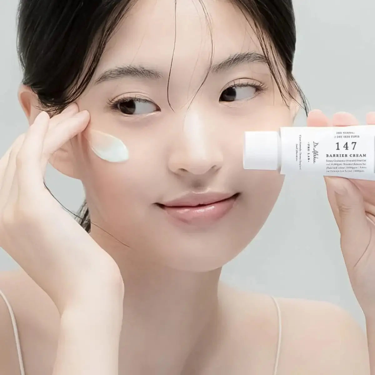 Woman applying cream to her face with a bottle labeled '147 Barrier Cream' on a neutral background