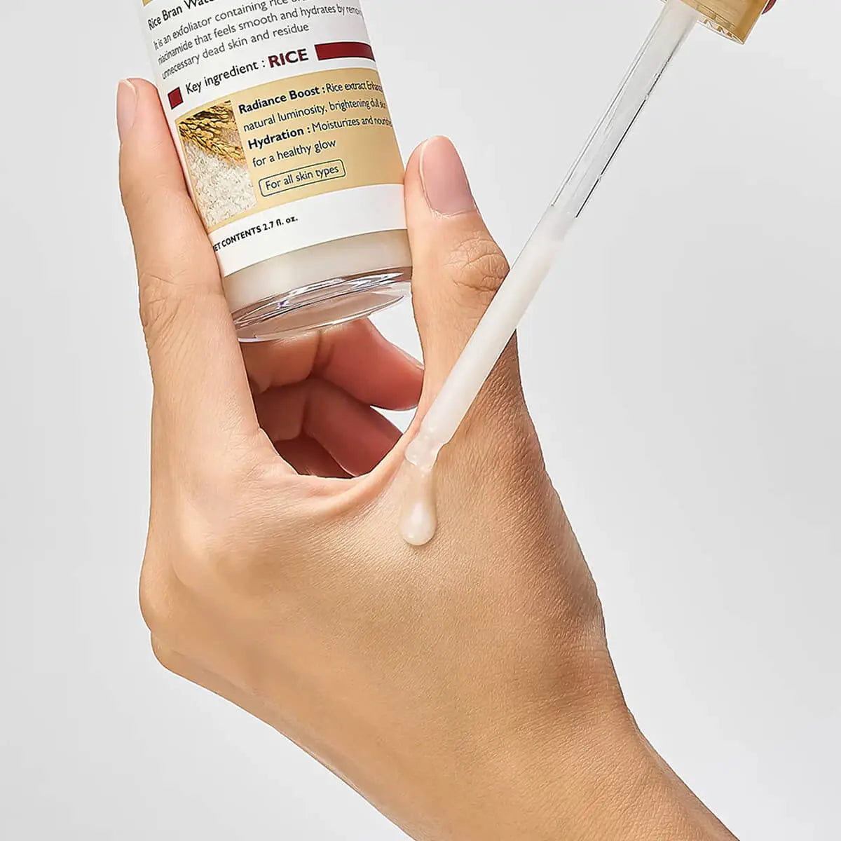 Hand holding a dropper with a skincare product and a bottle labeled 'Rice' on a light gray background