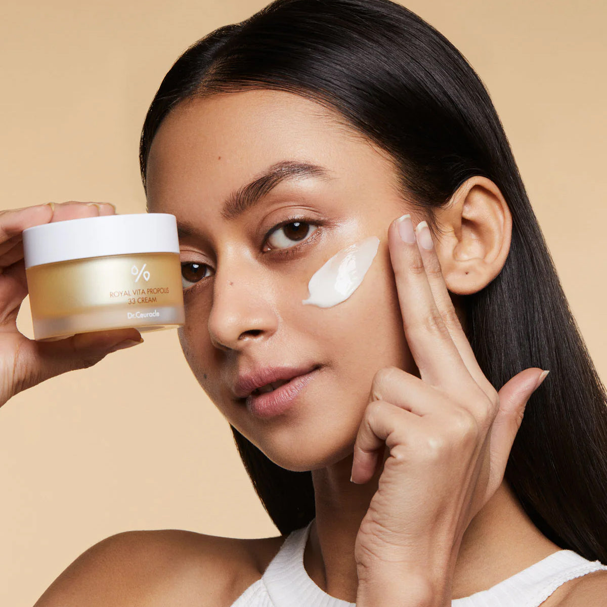 Woman applying cream to her face with a jar of cream in hand on a beige background