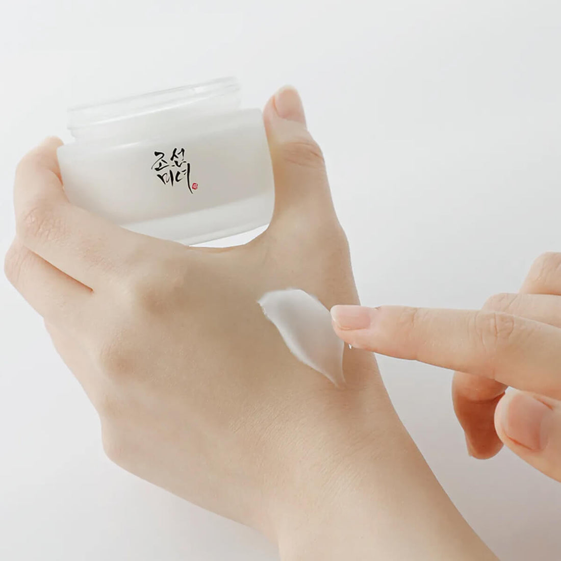 Hand applying cream from a jar with a visible brand logo on a white background