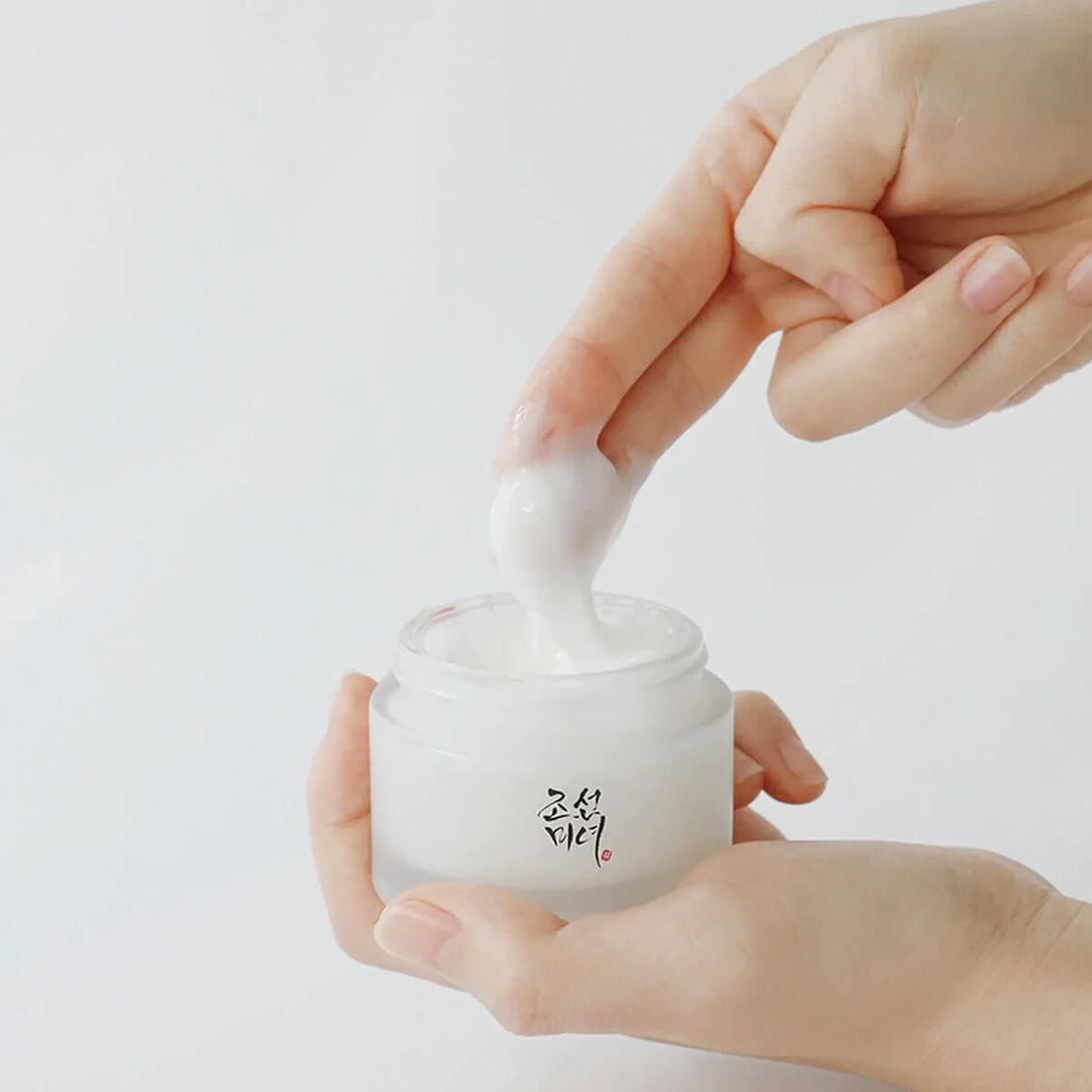 Hand holding a small jar with a white cream being scooped out on a light background