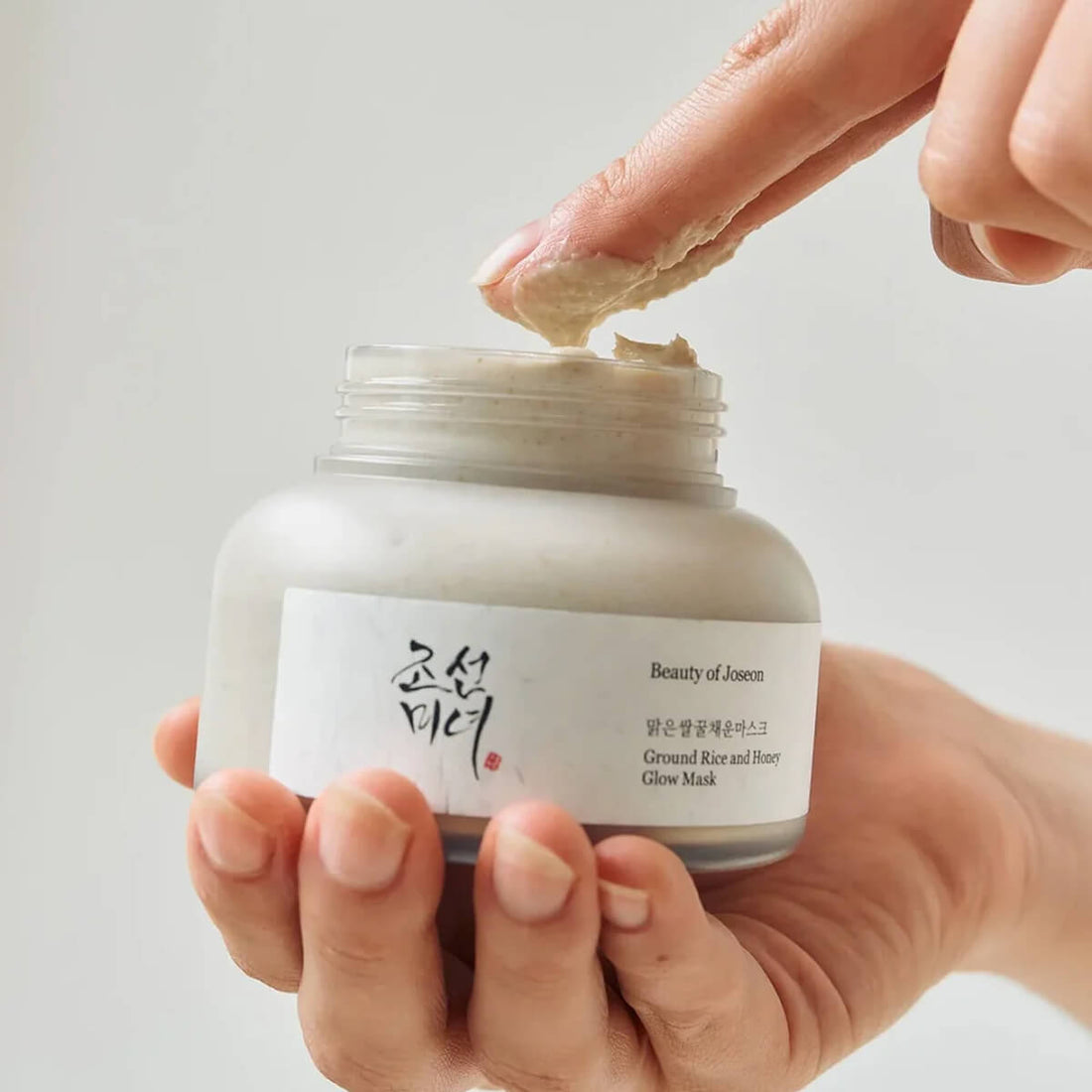 Hand applying cream from a jar labeled 'Beauty of Joseon' on a neutral background