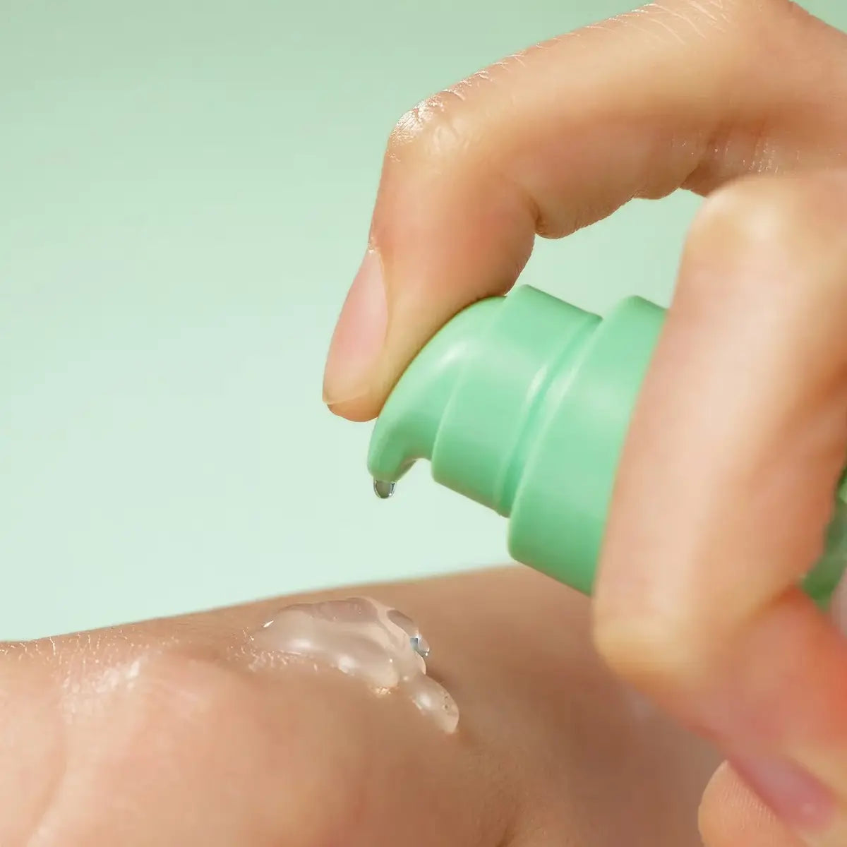 Hand applying a clear cream or lotion to skin with a green applicator against a light green background