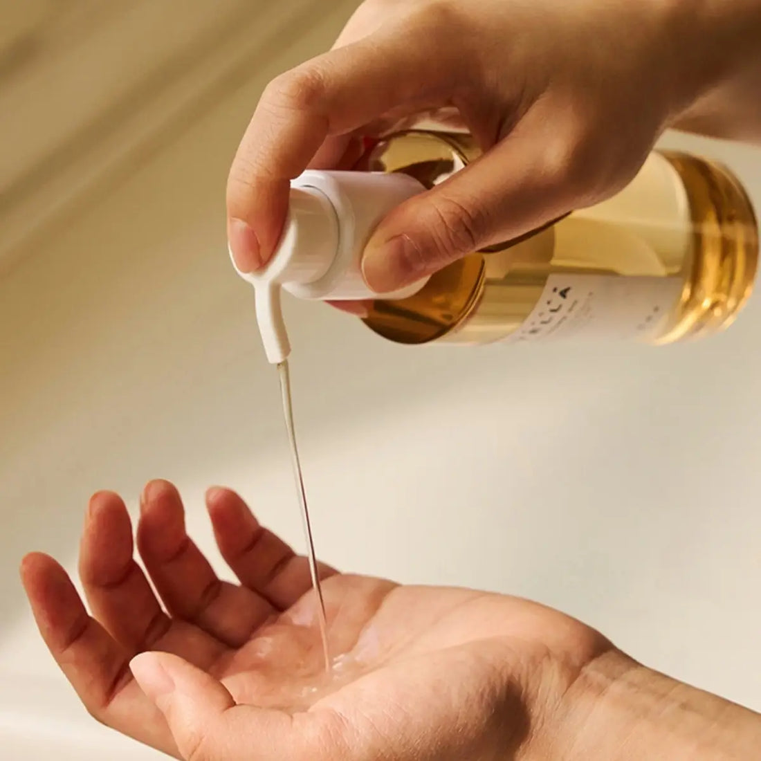 Hand pouring a clear liquid from a bottle into another hand against a neutral background