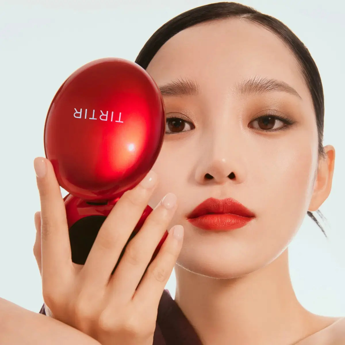 Woman holding a red compact mirror with 'TIRIR' branding against a light blue background