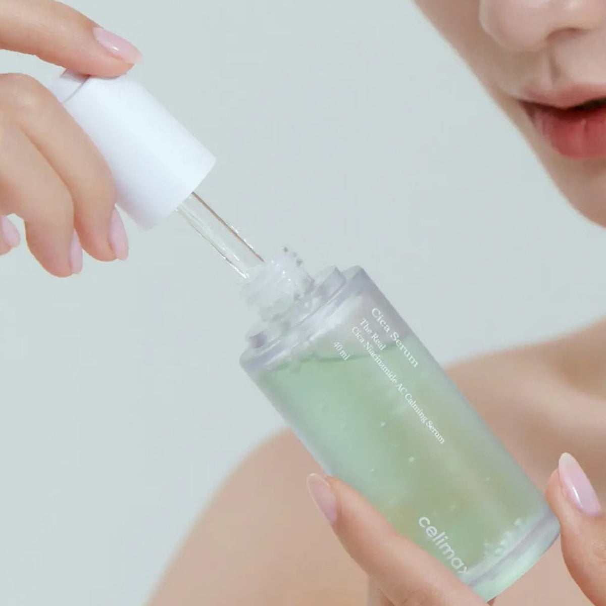Person holding a dropper bottle with a green liquid, likely serum or oil, against a neutral background.