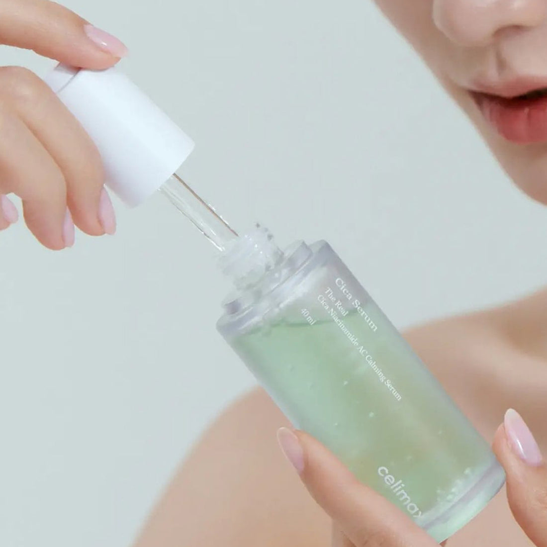 Person holding a dropper bottle with a green liquid, likely serum or oil, against a neutral background.