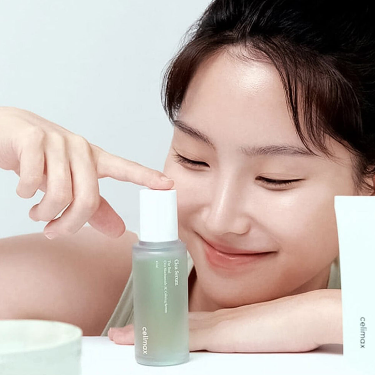 Woman applying a skincare product to her face with a bottle of Cetaphil serum in front of her.