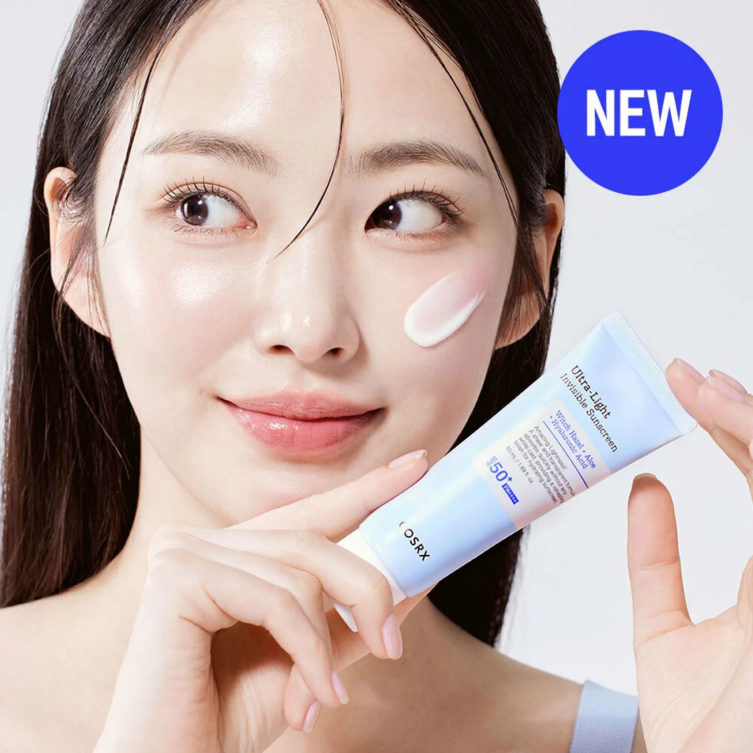 A person holding a tube of Cosrx Ultra Light Invisible Sunscreen SPF 50 PA++++. The product is applied to their cheek, and the tube is labeled 'NEW'.