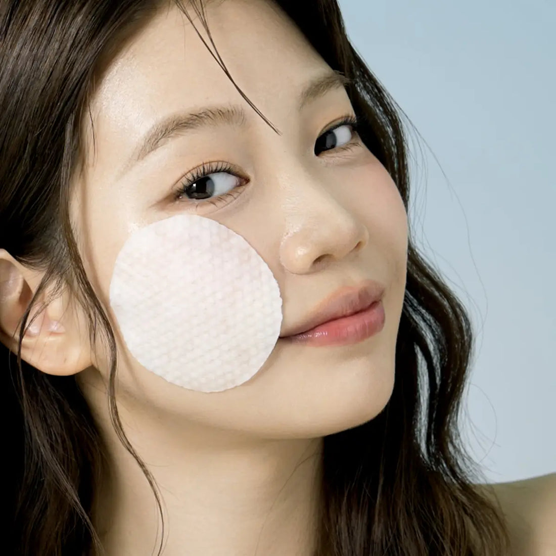 Woman applying a Eqqualberry Vegan Swimming Pool Toner Pad to her face against a light blue background