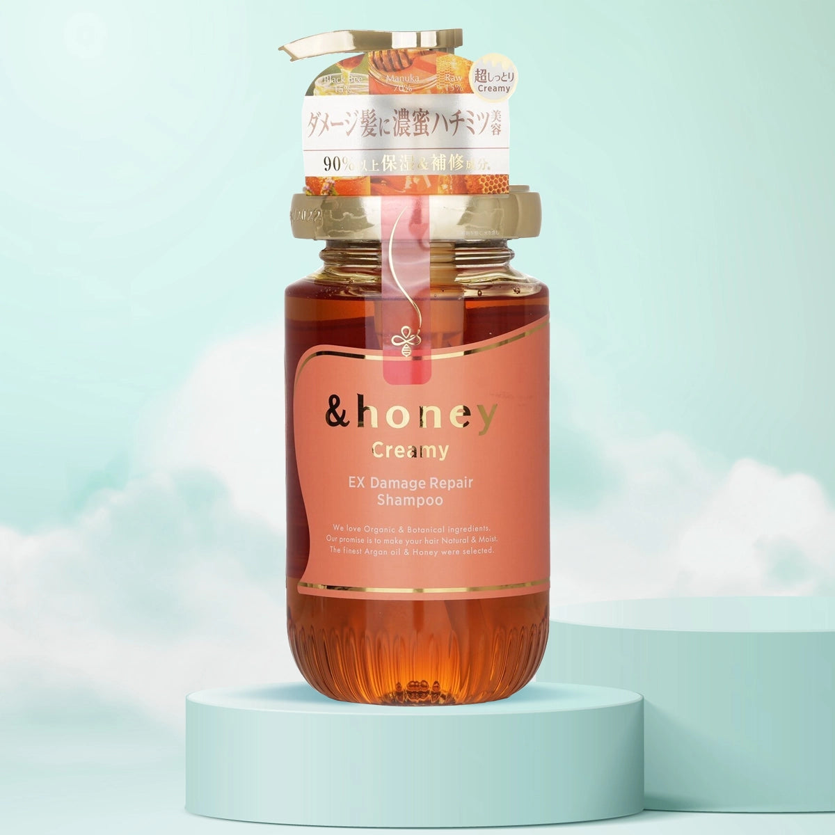 A bottle of &Honey Creamy EX Damage Repair Shampoo placed on a pedestal against a sky blue background, with its label and cap visible.