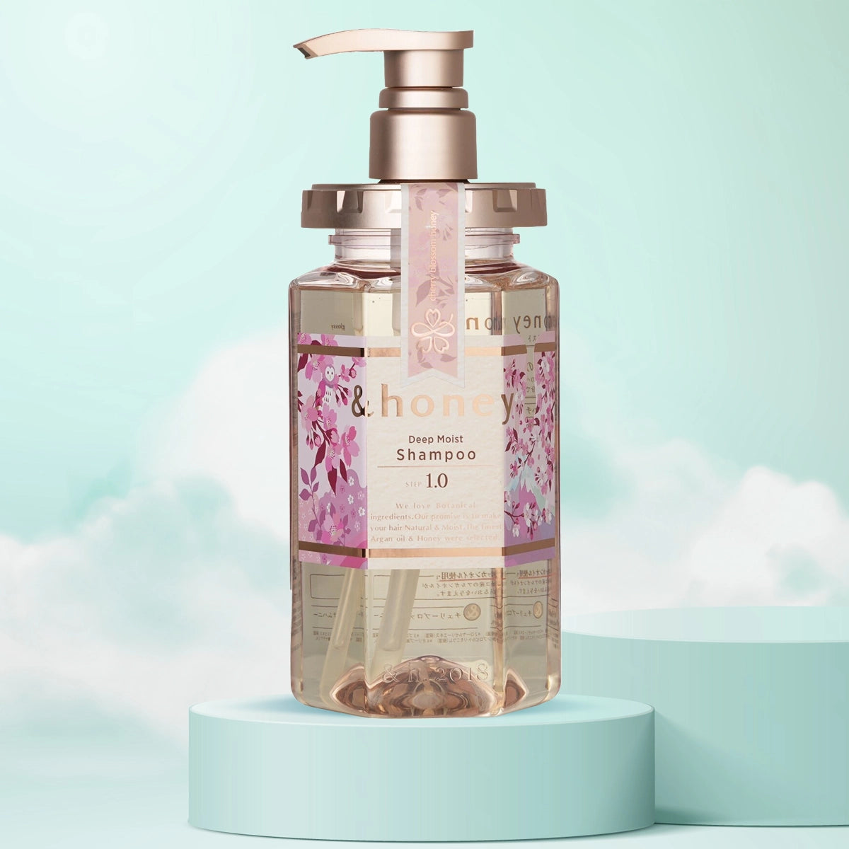 A pump bottle of &Honey Deep Moist Cherry Blossom Honey Shampoo with a pink label and floral design, presented on a teal pedestal with a light blue background.