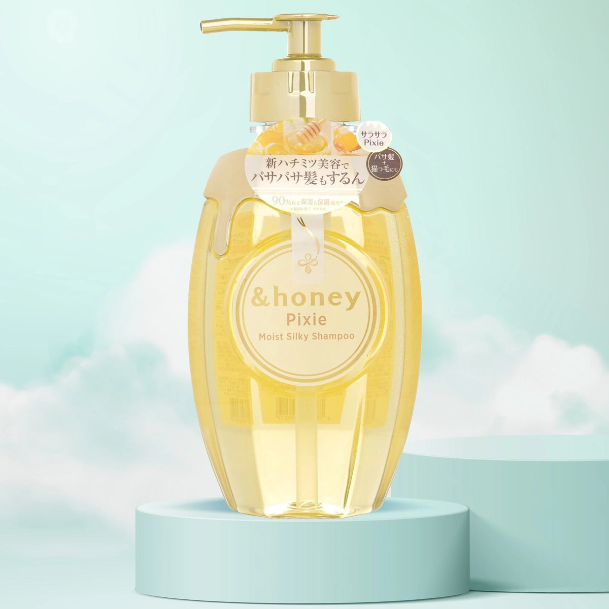 A clear pump bottle of &Honey Pixie Moist Silky Shampoo with a yellow liquid visible inside, placed on a light blue pedestal with a fluffy cloud background.