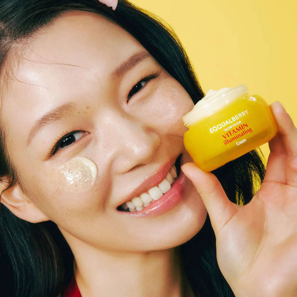 Woman applying cream to her face with a yellow skincare product in the foreground