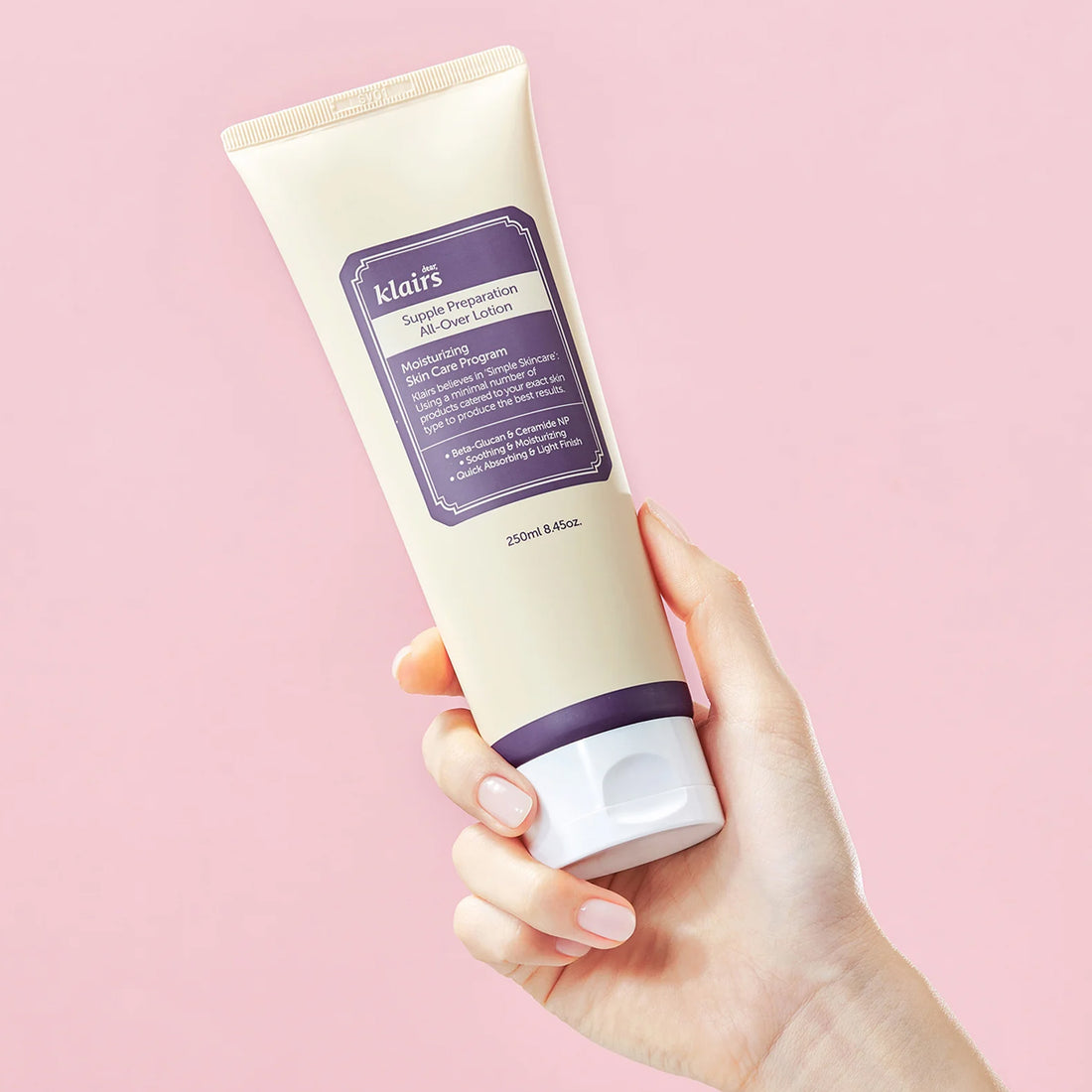 Hand holding a tube of Klairs Supple Preparation All Around Lotion against a pink background