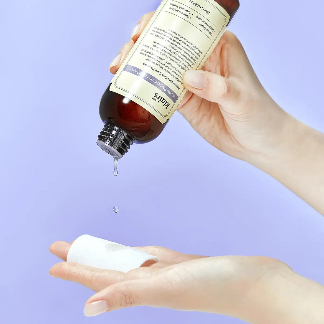 Hand holding a bottle of dear KLAIRS Supple Preparation Facial Toner droplet falling onto another hand against a light purple background